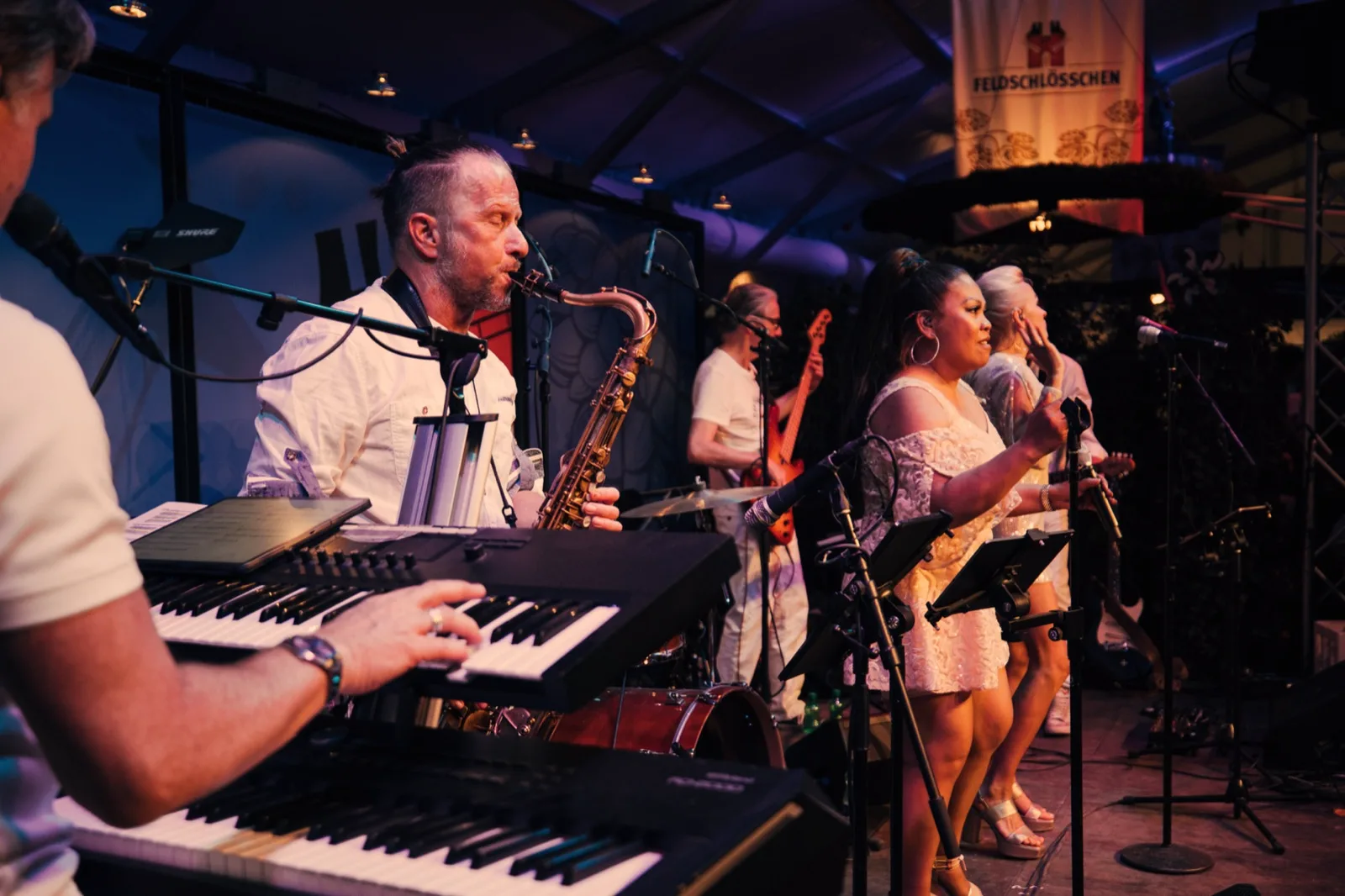 Abgedunkeltes Bühnen-Standbild einer Live-Show: Band mit Saxofon und Josephine Pee mit zweiter Sängerin rechts