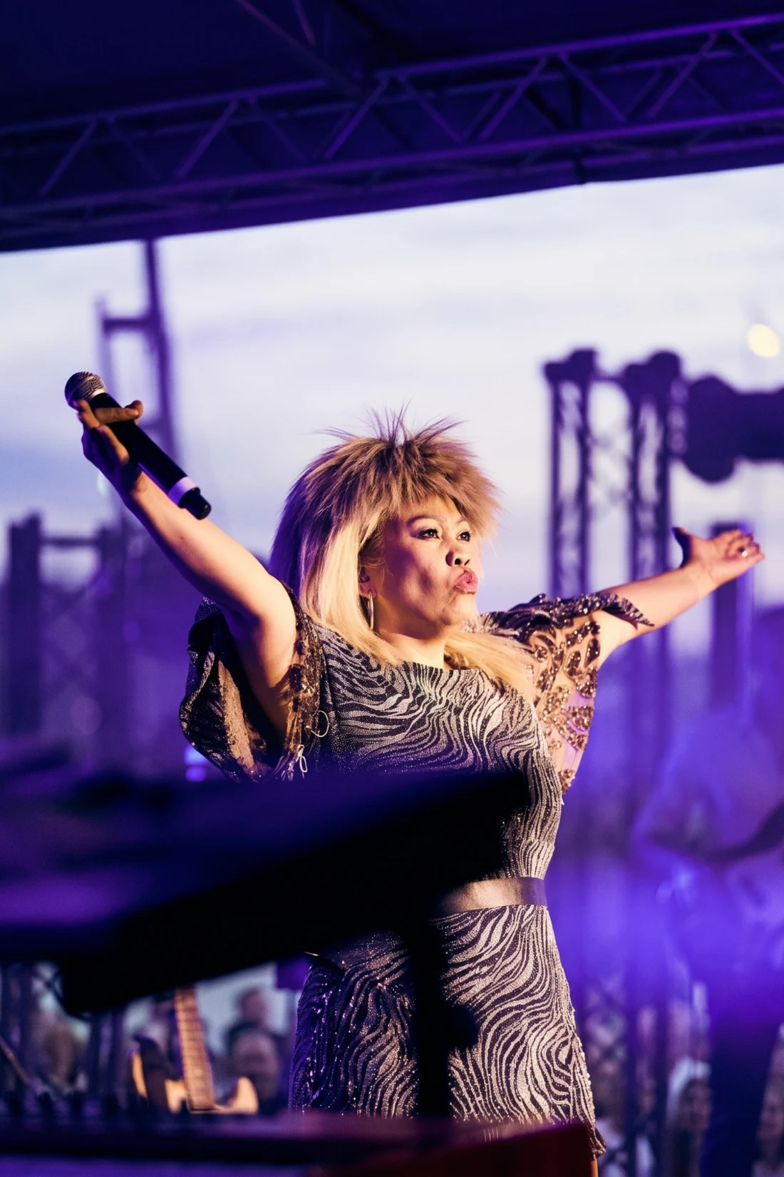 Josephine Pee als Tina Turner mit blonder Tina-Perücke, singend mit ausgestreckten Armen auf einer Open-Air-Bühne